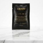 Um sachê preto de Amostra 15 em 1 - 15 ml está na vertical em uma superfície de mármore branco contra um fundo claro e liso, com texto dourado e branco exibindo detalhes do produto na embalagem.