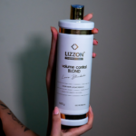 Uma pessoa segura um frasco branco grande do produto para cabelo Lizzon Professional Volume Control Blond com uma tampa dourada contra um fundo liso, mostrando a fórmula liso. O rótulo apresenta texto em preto e dourado com detalhes do produto.