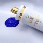 Um frasco branco e dourado de volume control blond (combo3x) repousa ao lado de uma mancha de creme roxo vibrante em uma superfície branca.