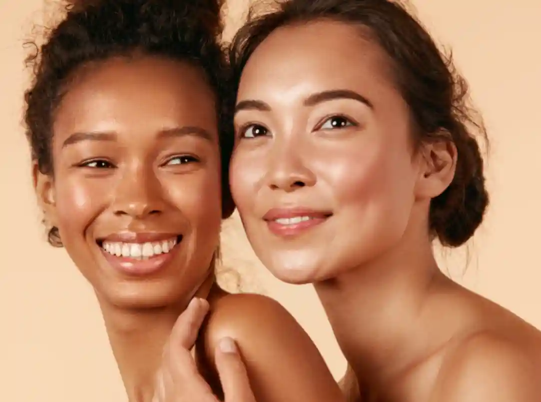 Duas mulheres com pele brilhante e saudável estão próximas uma da outra, sorrindo para a câmera contra um fundo bege neutro. Ambas estão com os cabelos presos para trás e parecem relaxadas e felizes, irradiando confiança com sua pele lisa e lisa.