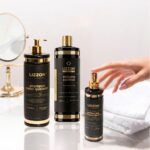 Três produtos de tratamento capilar Kit Liso Blindado estão dispostos em uma superfície branca ao lado de toalhas brancas dobradas, com uma mão alcançando um frasco e um espelho de mesa redondo visível ao fundo.