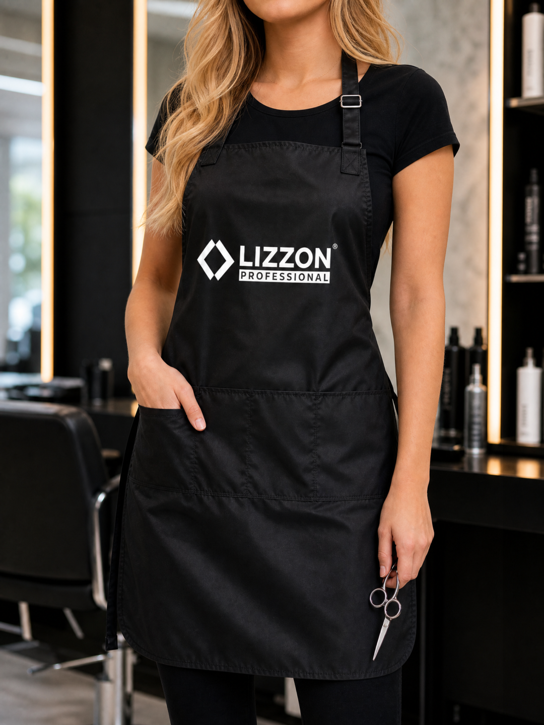 Vestindo o avental Avental Lizzon Professional, uma pessoa está em pé em um salão moderno segurando uma tesoura, com prateleiras de produtos para cabelos e cadeiras de salão ao fundo. Seu rosto permanece fora de cena.