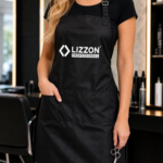 Vestindo o avental Avental Lizzon Professional, uma pessoa está em pé em um salão moderno segurando uma tesoura, com prateleiras de produtos para cabelos e cadeiras de salão ao fundo. Seu rosto permanece fora de cena.