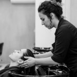 Um cabeleireiro usando um avental de liso lava gentilmente o cabelo de uma cliente em um lavatório de salão. A cliente está recostada com os olhos fechados em um ambiente moderno, com prateleiras e produtos para cabelo visíveis ao fundo.