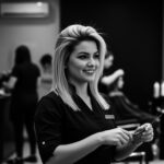 Um cabeleireiro com cabelo liso e loiro na altura dos ombros (liso) segura um pente em um salão moderno, sorrindo. Outros clientes e estilistas são visíveis ao fundo, com prateleiras de produtos para cabelo, incluindo Lizzon, ao longo da parede.