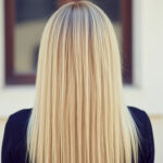 Uma pessoa com cabelo liso, louro e comprido é mostrada de costas, em pé em frente a uma janela em uma parede de cor clara. O indivíduo está vestindo uma blusa escura.