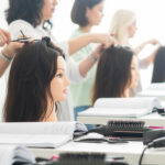 Pessoas praticam técnicas de modelagem de cabelo liso em cabeças de manequins em uma sala de aula. Escovas de cabelo, secadores de cabelo e cadernos abertos estão sobre as mesas, e várias mulheres se concentram em seu trabalho. A sala é bem iluminada.