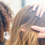 Uma pessoa examina o couro cabeludo e o cabelo de outra pessoa com cabelo liso, liso e castanho claro. Ao fundo, vê-se outra pessoa com cabelos cacheados e castanho-escuros. O cenário parece ser um salão de cabeleireiro lizzon ou ambiente semelhante.