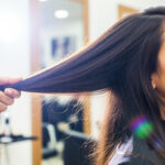 Uma mulher senta-se em uma cadeira de salão enquanto um estilista examina uma seção de seu longo cabelo castanho liso. O fundo do salão está suavemente desfocado.