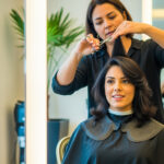 Um cabeleireiro corta o cabelo liso, na altura dos ombros, de uma mulher em um salão moderno. A mulher está sentada em uma cadeira, usando uma capa preta de salão e olhando para frente, enquanto o cabeleireiro trabalha com uma tesoura atrás dela.