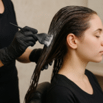 Uma pessoa usando luvas pretas aplica tinta de cabelo com um pincel no cabelo molhado de uma mulher em um salão de beleza. A mulher, sentada em uma cadeira com os olhos fechados, está vestindo uma camisa preta.