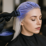 Uma pessoa com cabelos longos, lisos e loiros está sentada em uma cadeira de salão com os olhos fechados enquanto alguém usando luvas pretas aplica tinta roxa em suas raízes usando um pincel.