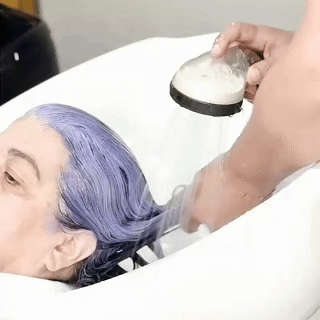 O cabelo de uma pessoa, pintado com tinta roxa, está sendo enxaguado com água por outra pessoa na pia de um salão de beleza.