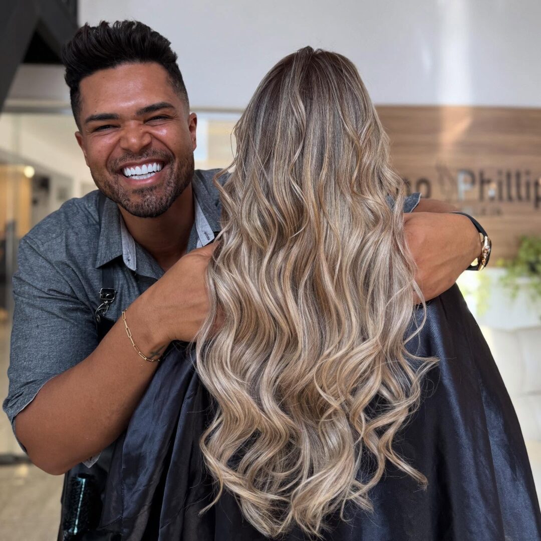 Um cabeleireiro sorridente está atrás de uma cliente com cabelos longos, ondulados e com mechas loiras, modelados com Pó Ultra Rápido Descolorante PLEX 1kg (Combo 2x), em um salão de beleza. A cliente está de costas para a câmera.