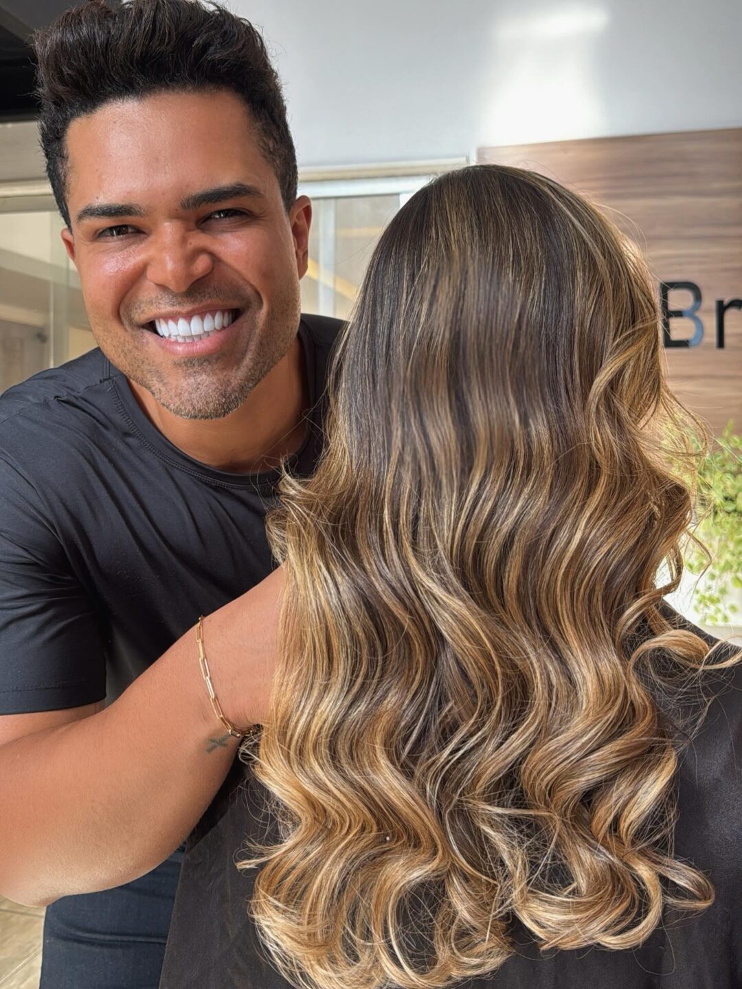 Um homem sorridente está atrás de uma mulher com cabelos longos, ondulados e realçados, criados com Pó Ultra Rápido Descolorante PLEX 1kg (Combo 2x). O foco está em seu cabelo luminoso, em um ambiente interno com a luz do sol passando por uma janela.