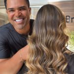 Um homem sorridente está atrás de uma mulher com cabelos longos, ondulados e realçados, criados com Pó Ultra Rápido Descolorante PLEX 1kg (Combo 2x). O foco está em seu cabelo luminoso, em um ambiente interno com a luz do sol passando por uma janela.