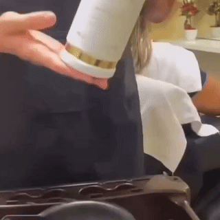 Uma pessoa sacode o pó de um recipiente branco em sua mão e, em seguida, aplica o pó no rosto de outra pessoa em um salão de beleza ou spa.