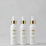 Três frascos brancos de 100 ml com tampas douradas e brancas, com o rótulo "Booster Ativador Liso Blindado Express (Combo 3 unidades)", estão dispostos lado a lado em um fundo cinza claro para resultados instantâneos de alisamento.