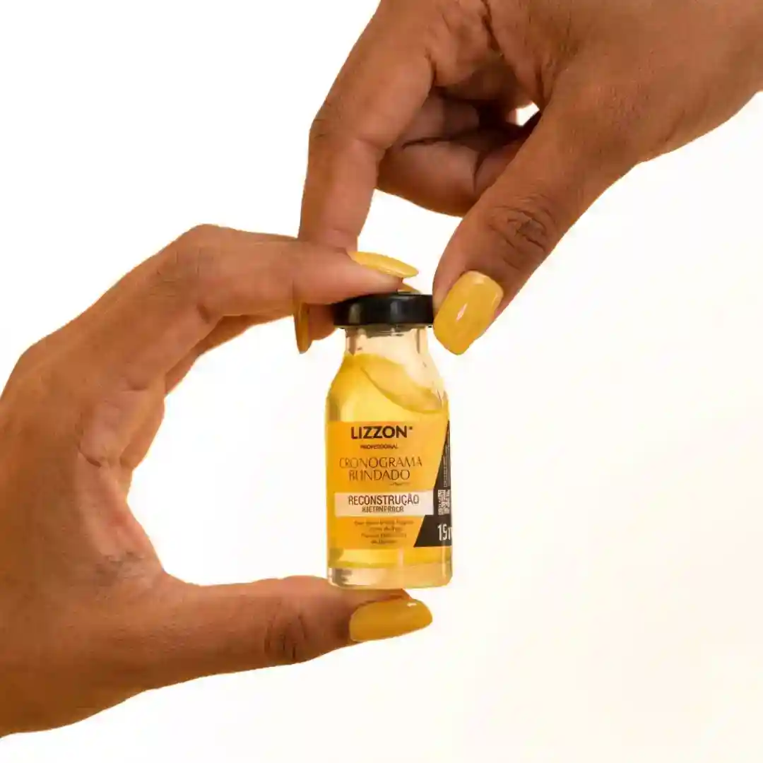 Duas mãos com unhas pintadas de amarelo seguram um pequeno frasco de vidro contendo um líquido amarelo do Kit Power Dose. O rótulo do frasco diz "Lizzon Cronograma Blindado Reconstrução". O plano de fundo é branco.