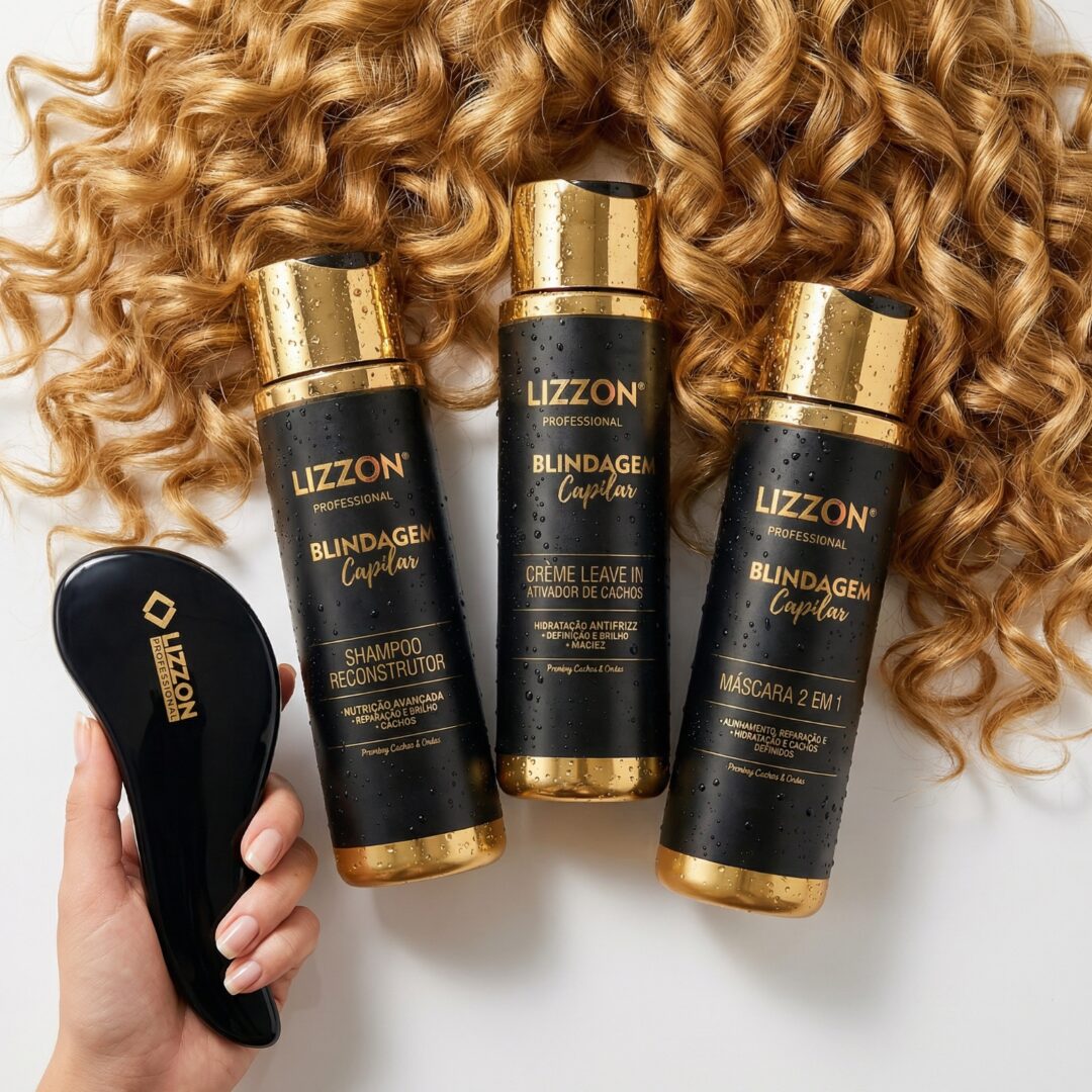 Três frascos de kit cachos blindados (cuidados domésticos) - xampu, creme sem enxágue e máscara 2 em 1 - além de uma escova preta para desembaraçar estão dispostos em um cabelo cacheado louro mel. Uma mão é mostrada segurando a escova.