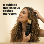 Uma mulher de cabelos longos e cacheados sorri com os olhos fechados, segurando um frasco de Ativador de Cachos: Creme Leave In Blindagem Capilar. A imagem apresenta um texto em português que promove o cuidado com os cabelos cacheados.