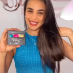 Uma mulher sorridente, com longos cabelos escuros e um top azul sem mangas segura a caixa do Kit Power Dose Cronograma Blindado (Combo 3 unidades), exibindo seus três recipientes. O fundo está desfocado.