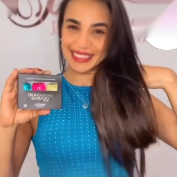 Uma mulher sorridente, com longos cabelos escuros e um top azul sem mangas segura a caixa do Kit Power Dose Cronograma Blindado (Combo 3 unidades), exibindo seus três recipientes. O fundo está desfocado.