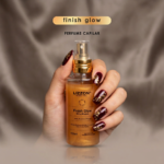 Uma mão com nail art em vermelho escuro e dourado segura um frasco de Lizzon Finish Glow Perfume Capilar em um fundo bege acetinado. O texto acima do frasco diz
