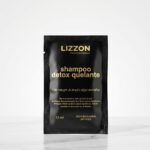 Um sachê preto com o rótulo "Amostra Shampoo Detox Quelante - 15 ml", com texto em dourado e branco, está na vertical em uma superfície de mármore branco contra um fundo branco liso.