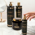 Quatro frascos pretos e dourados do Kit Liso Blindado (cópia) estão dispostos em uma superfície branca perto de um espelho redondo. Uma mão está prestes a borrifar um dos produtos: xampu, controle de volume, máscara capilar ou spray térmico.