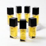 Seis frascos de líquido amarelo rotulados como Ouro Inka by Lizzon estão dispostos em um círculo em uma superfície branca, com três frascos elevados na parte de trás e três na frente. Cada frasco tem uma tampa preta e um corpo retangular transparente.