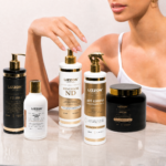 Uma mulher vestindo uma regata branca exibe o Kit Tratamento Lizzon 1, que inclui seis produtos para os cabelos dispostos sobre uma superfície de mármore. Sua mão está erguida, com os dedos levemente curvados, acima dos frascos, que se destacam por suas embalagens nas cores branca, preta e dourada.