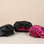 Três gorros de cetim da TOUCA DE CETIM DUPLA FACE BIG, nas cores preta e rosa escuro, estão dispostos sobre uma superfície clara com fundo neutro. Os gorros apresentam bordas franzidas e um tecido brilhante e liso.
