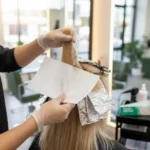 Usando luvas, uma cabeleireira aplica tintura em cabelos loiros usando folhas de alumínio e o Papel Para Mechas Luzes 11x47cm Padrão 30 Folhas em um salão de beleza, com cadeiras e equipamentos do salão visíveis no fundo bem iluminado.