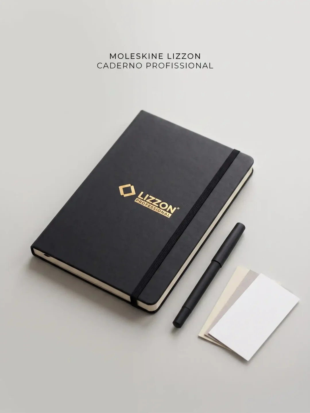 Um caderno profissional Lizzon preto com uma faixa elástica, uma caneta preta e dois cartões em branco estão dispostos sobre uma superfície cinza-clara. O texto acima diz: Moleskine Lizzon Caderno Profissional..