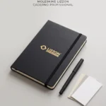 Um caderno profissional Lizzon preto com uma faixa elástica, uma caneta preta e dois cartões em branco estão dispostos sobre uma superfície cinza-clara. O texto acima diz: Moleskine Lizzon Caderno Profissional..